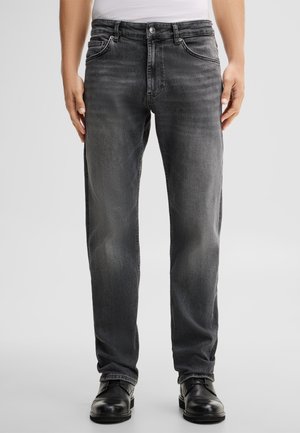 Man wearing straight-fit dark gray jeans and black leather shoes standing on a plain light background.