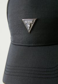 Czarna materiałowa czapka z trójkątną srebrną naszywką z napisem "GUESS USA" i znakiem zapytania. Gładka tekstura z przeszyciami na daszku.