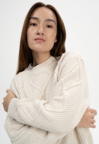 Cremfarbener Cable-Knit-Pullover mit einem gerippten Kragen und geometrischen Mustern. Weicher, strukturierter Stoff mit lockerer Passform und langen Ärmeln.