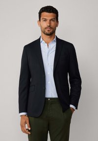 Blazer navy avec des épaules structurées, deux poches avant et une fermeture à un bouton, porté sur une chemise col bleu clair et un pantalon vert olive.