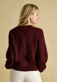 Bordový pletený sveter s textúrovaným vzorom a diamantom, s rebrovanými manžetami a lemom a voľným strihom, nosený s svetlými džínsami.
