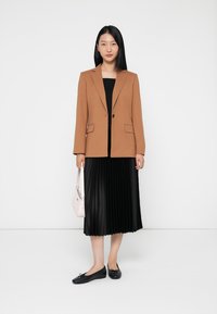 Blazer marrone con un solo bottone, abbinato a una gonna plissettata nera. Modella una borsa rosa e delle ballerine nere, su uno sfondo bianco.
