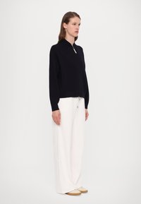 Marineblå zip-up genser med høy krage, myk tekstur, avslappet passform, parret med vide hvite bukser og lyse sko. Enkelt design.