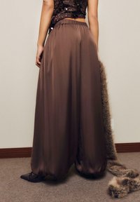 Brązowe satynowe spodnie szerokie z elastycznym paskiem w taliach, o gładkiej fakturze i zwężających się mankietach, zestawione z czarnymi, spiczastymi butami.