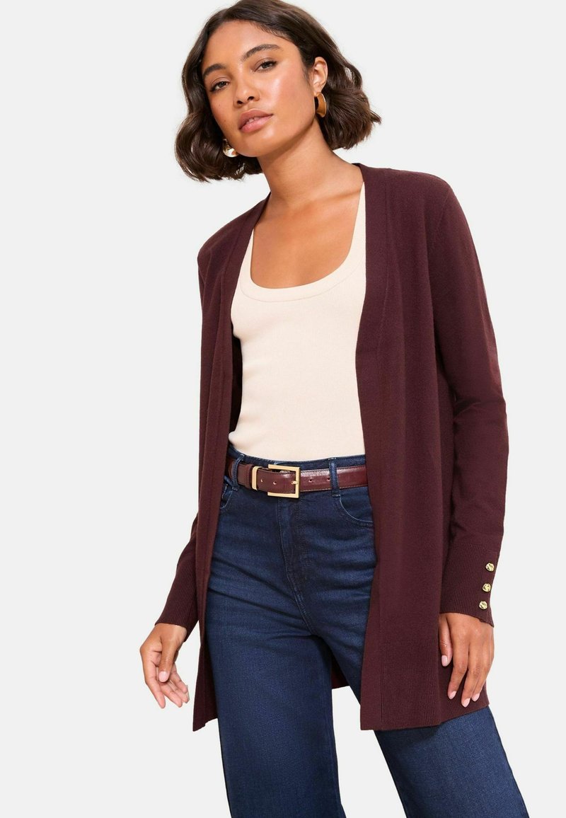 Gilet bordeaux avec une ouverture frontale, des manches longues et des accents de boutons dorés. Associé avec un top en crème et un jean sombre, affichant une coupe décontractée.