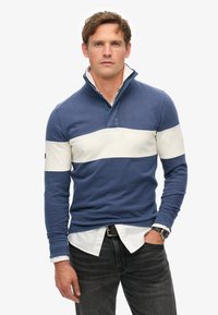 Blauwe polo met lange mouwen en een witte horizontale strook over de borst. Zachte textuur, kraag met kwart-rits, gedragen over een wit shirt.