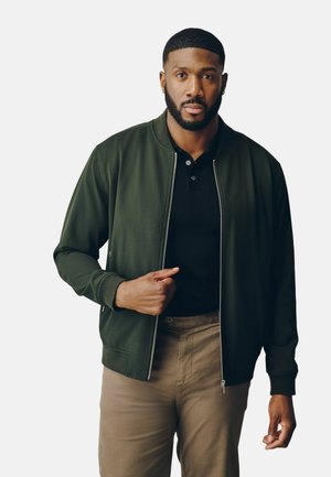Mann mit kurzen Haaren und Bart trägt eine grüne Zip-up-Jacke über einem schwarzen Hemd und beigefarbenen Hosen, posiert vor einfarbig hellem Hintergrund.