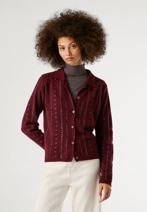 Cardigan en tricot bordeaux avec un col, présentant des motifs verticaux et une fermeture à boutons, superposé à un col roulé rayé.