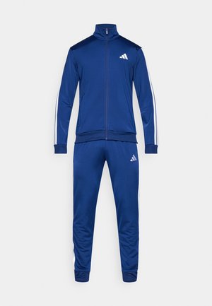 Chándal azul con cremallera completa, con rayas blancas en las mangas y los pantalones, y un pequeño logo blanco de Adidas en el pecho y el muslo izquierdo.