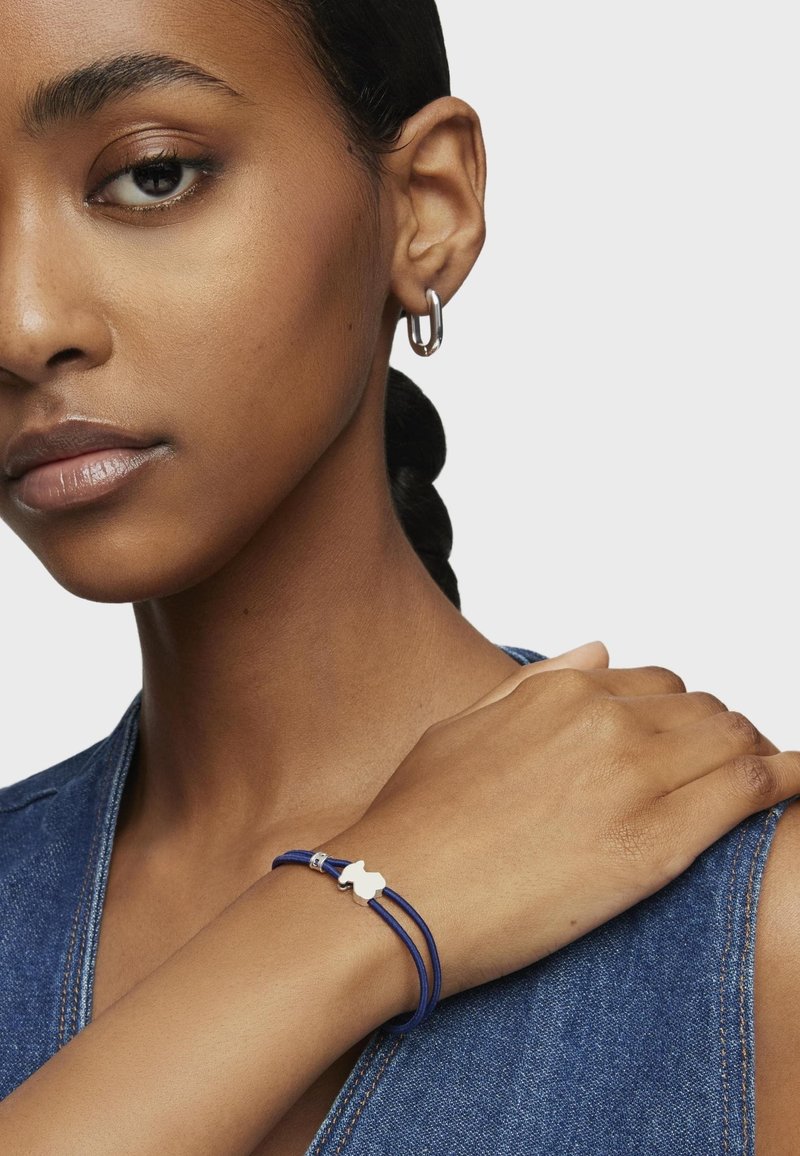 Bracciale blu in corda con un accento in argento, indossato su un polso. Il modello indossa una maglietta di denim e un orecchino a cerchio, con un’acconciatura semplice e intrecciata.