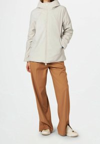 Veste beige clair zippée avec capuche, présentant une texture lisse et deux poches avant. Associée à un pantalon à jambes larges marron et des baskets.