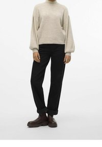 Personne portant un pull beige avec des manches ballon, un pantalon noir retroussé et des bottines marron foncé, debout devant un fond uni.