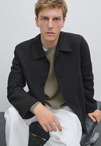 Sort uldfrakke med krave, knaplukning og brystnæse detaljer, båret over en tekstureret beige sweater og hvide bukser.