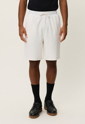 Pantaloncini bianchi in cotone con vita elasticizzata e coulisse; abbinati a calzini neri e scarpe nere; texture liscia e design casual.