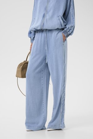 Osoba nosząca jasnoniebieskie szerokie dżinsowe spodnie i dopasowaną kurtkę, trzymająca małą brązową torebkę, z jedną ręką w kieszeni.