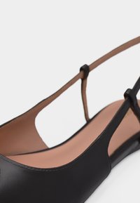 Μαύρο δερμάτινο φλατ παπούτσι με λουράκι στο πίσω μέρος, με ταμπά μαλακό εσωτερικό πέλμα και λεπτό ρυθμιζόμενο λουράκι στον αστράγαλο, σε λευκό φόντο.