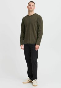 Langärmliges, olivgrünes Henley-Shirt mit drei Knöpfen, kombiniert mit schwarzen Jeans und beigen Sneakers mit braunen Streifen. Einfaches Design.