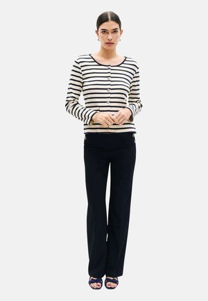 Femme portant un cardigan à boutons rayé bleu marine et blanc, un pantalon noir à jambes larges, et des talons ouverts bleu marine, debout les mains jointes.