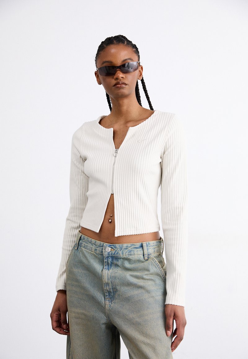 White ribbed knit cropped jacket with long sleeves and a front zipper. Paired with faded blue high-waisted jeans. Small logo detail on the jacket.
