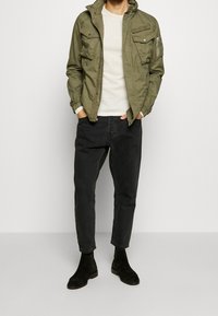 Olivgrön zip-up-jacka med flera fickor, cremefärgad tröja, svarta avsmalnande jeans och svarta ankelstövlar. Slätt tyg och strukturerad design.