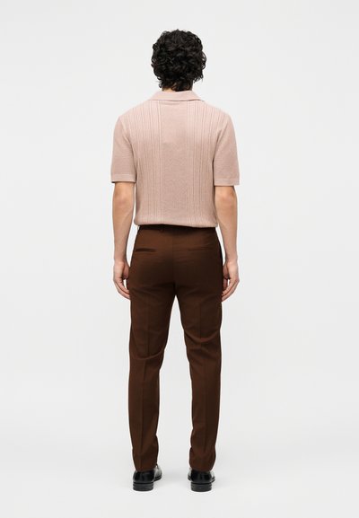 Άνδρας που στέκεται με την πλάτη, φοράει ένα μπεζ πλεκτό πουλόβερ με κοντά μανίκια, καφέ ραμμένο παντελόνι και μαύρα παπούτσια, με λευκό φόντο.