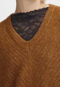 Sweter z dzianiny w kolorze rdzy z dekoltem w serek, zdobiony zygzakowatym wzorem i delikatnym połyskiem. Nałożony na czarną koronkową bluzkę.