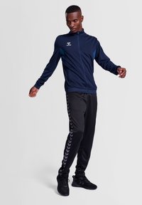 Mörkblå zip-jacka med ljusblå detaljer och Hummel-logotyp; matchad med svarta byxor som har ett grått chevronmönster längs sidorna.