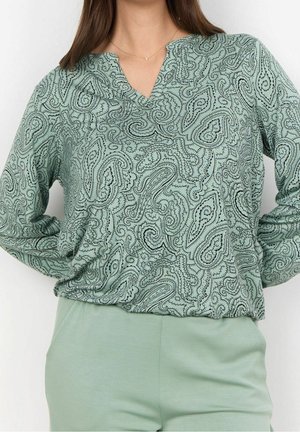 Femme portant un haut à manches longues vert clair à motif cachemire avec un col en V et un pantalon uni vert clair, mains sur les hanches.