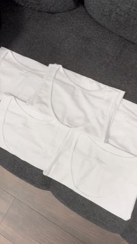 Four white cotton tank tops stacked, featuring a rounded neckline and wide shoulder straps. The fabric appears smooth and lightweight.