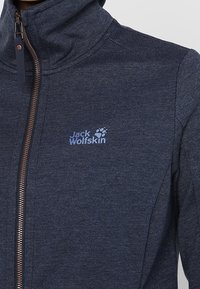 Námořnická modrá fleecová bunda s vysokým límcem, vybavená bronzovým zipem a světle modrým logem. Měkká textura s sportovním designem.