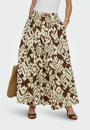 Donna che indossa una gonna lunga marrone e bianca con tasche, abbinata a sandali con tacco marroni e una borsa intrecciata.