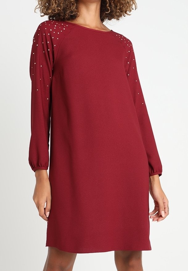 Femme portant une robe rouge longue jusqu'aux genoux avec des manches longues et de petites clous décoratifs sur les épaules et le haut des manches.