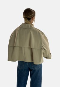 Giacca beige corta con design a strati sul retro, ampio colletto e maniche lunghe, realizzata in un tessuto liscio, abbinata a jeans in denim blu.