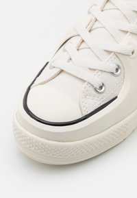 Gros plan sur une chaussure en toile blanche avec des lacets blancs, des œillets métalliques, un embout en caoutchouc et une semelle texturée blanc cassé avec un liseré noir.