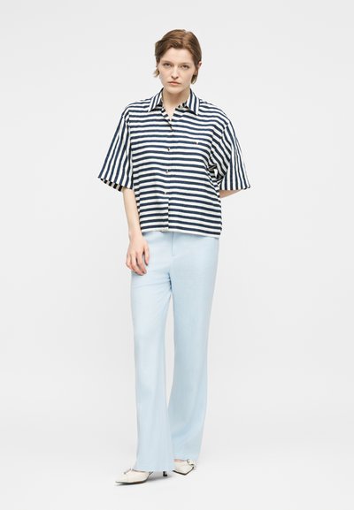 Γυναίκα που στέκεται φορώντας ανοιχτό μπλε παντελόνι, άσπρα μυτερά παπούτσια και μια χαλαρή κοντομάνικη μπλούζα με κουμπιά σε σκούρο μπλε και λευκές ρίγες.