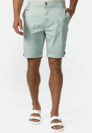 Mann trägt hellgrüne Shorts mit umgeschlagenem Saum und braunen Knöpfen, weiße Sandalen mit doppeltem Riemen, stehend mit Händen in den Taschen vor weißem Hintergrund.