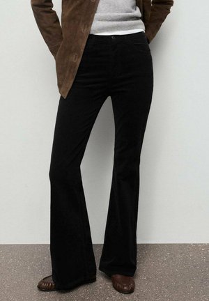 Pantalones negros acampanados en tela suave, diseñados con una cintura de altura media y bolsillos frontales. Combinados con un suéter gris claro y una chaqueta de ante marrón.