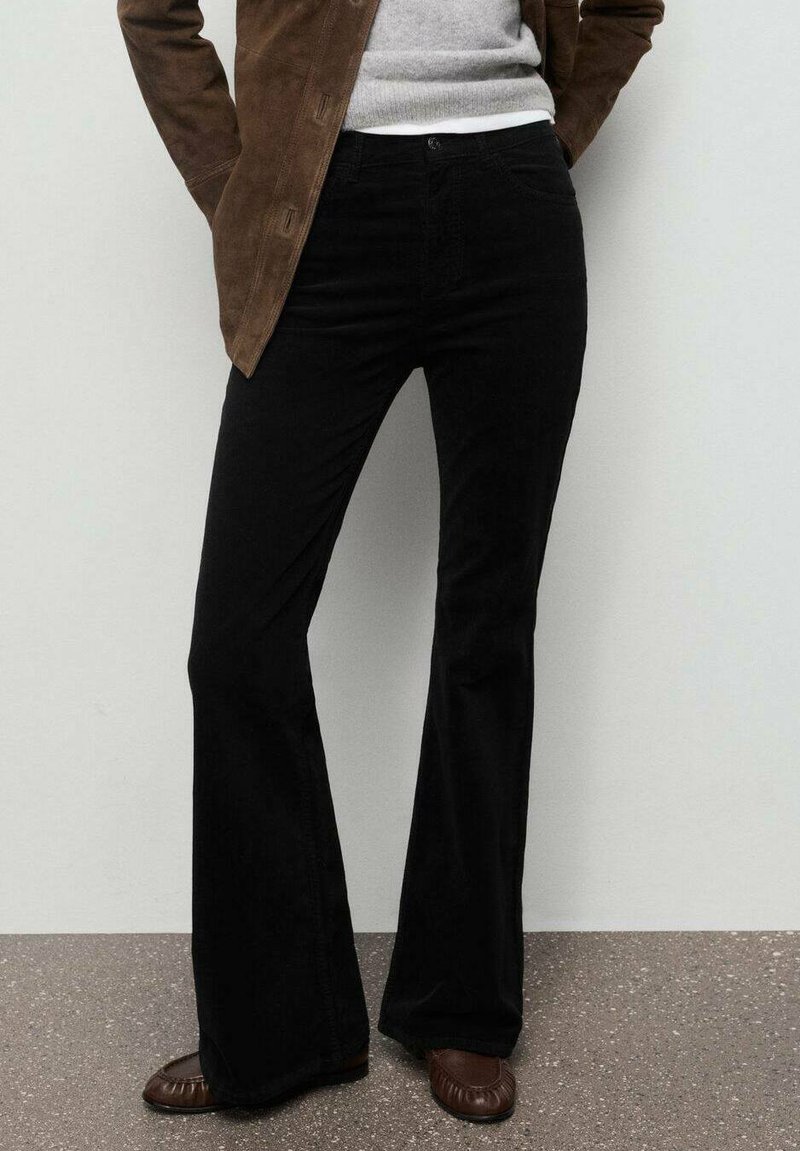 Pantalones negros acampanados en tela suave, diseñados con una cintura de altura media y bolsillos frontales. Combinados con un suéter gris claro y una chaqueta de ante marrón.