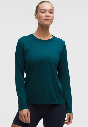Vrouw draagt een langarmige teal trui met ronde hals en zwarte strakke broek, staand tegen een effen lichtgrijze achtergrond.