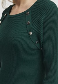 Top verde a coste con scollatura ampia, bottoni metallici decorativi sulla spalla e maniche lunghe. Tessuto testurizzato.