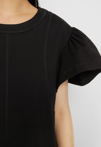 Haut noir avec des détails de coutures verticales et des manches courtes bouffantes sur une personne à la peau claire sur un fond gris uni.
