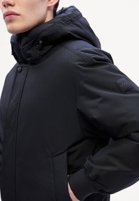 Veste noire isolée avec un col montant et une capuche ajustable. Comprend des poignets élastiques, des poches latérales, et un petit patch logo sur la manche.