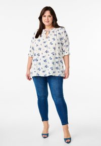 Blusa a fiori color crema con fiori blu, caratterizzata da un collo con apertura a chiave e lacci. Indossata con jeans skinny blu e sandali aperti.