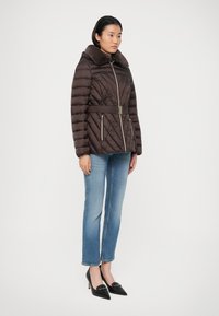 Veste matelassée brune avec un col en fausse fourrure, taille ceinturée et coutures diagonales. Associée à un jean bleu et des escarpins noirs à bout pointu.