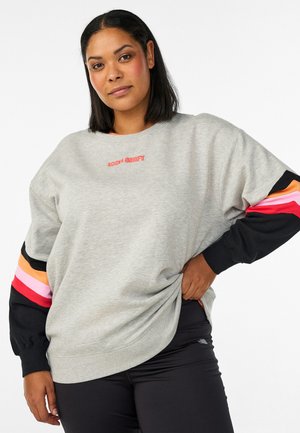 Žena s rovnými čiernymi vlasmi, oblečená v sivom tričku s viacfarebnými pruhovanými rukávmi a čiernymi nohavicami, pózujúca s rukou na bokoch.