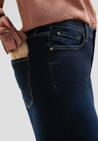 Tmavě modré denimové džíny s úzkým střihem, viditelné prošívání v kontrastní zlaté barvě a detail přední kapsy. Tkanina vypadá mírně strukturovaně.