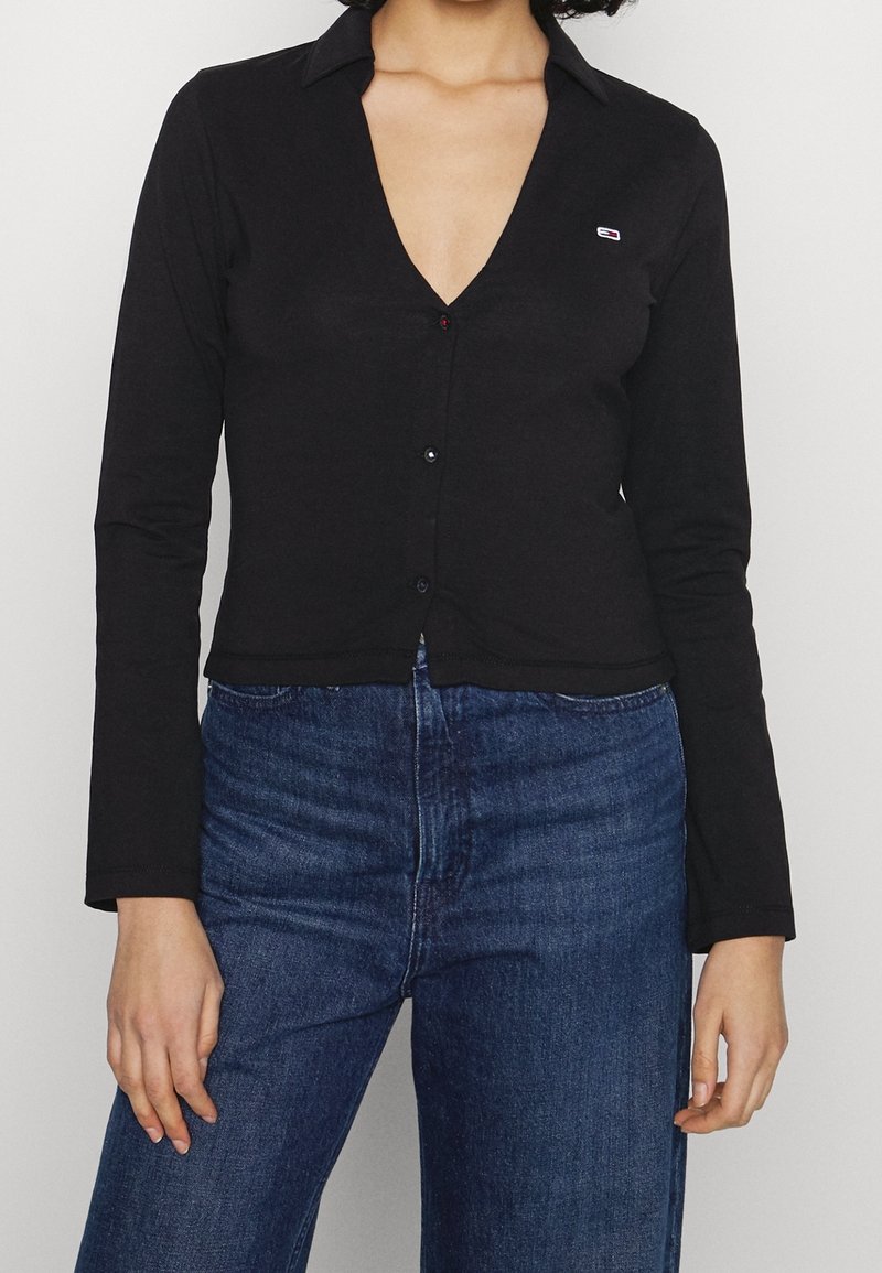 Black long-sleeve shirt with a deep V-neck, button closure, and subtle logo on the upper left chest. Soft, smooth fabric with a fitted design.