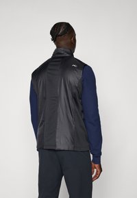 Čierny bezrukávový sveter s vysokým golierom, vyrobený z hladkého syntetického materiálu, ktorý má textúrovanú zadnú časť a logo na pleci.