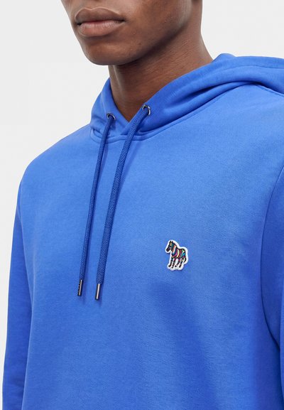 Homme portant un sweat à capuche bleu avec des cordons et un petit patch zèbre multicolore sur la poitrine sur un fond uni.