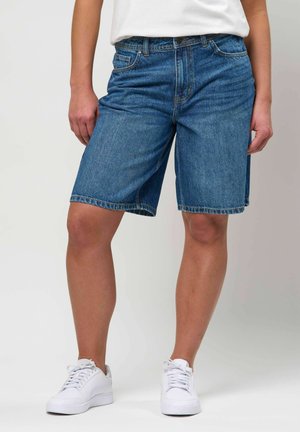 Osoba ubrana w niebieskie szorty z denimu sięgające do połowy uda, białe trampki i białą koszulę, stojąca na tle jednolitego jasnego tła.